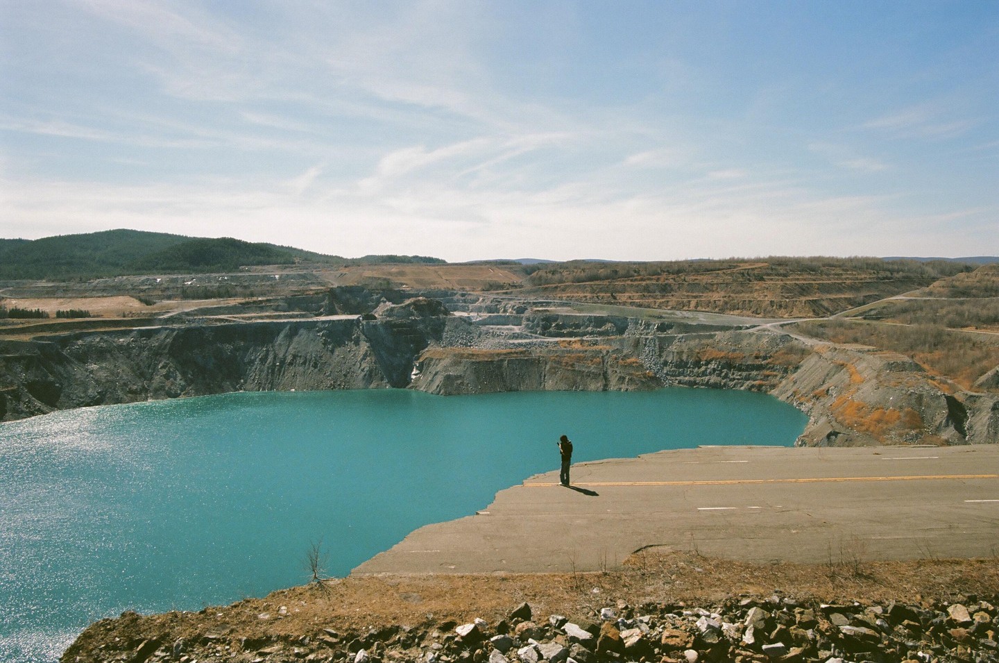 A person stands on a broken highway overlooking a large turquoise lake surrounded by a quarry.