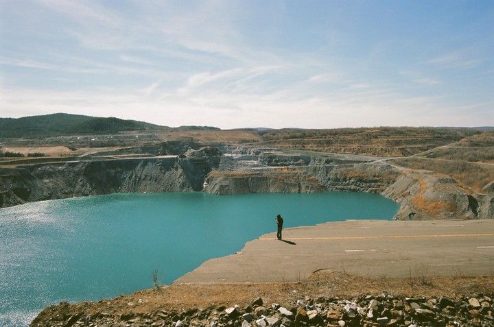 A person stands on a broken highway overlooking a large turquoise lake surrounded by a quarry.