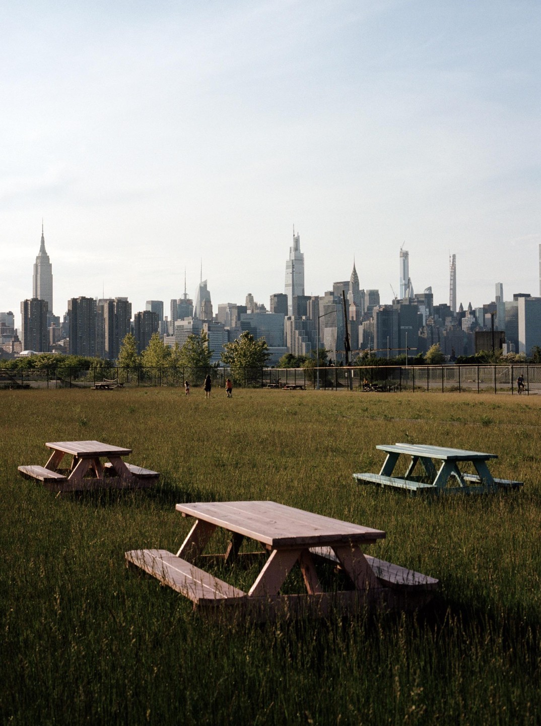 Three picnic tables sit in a grassy field with a city skyline in the background.