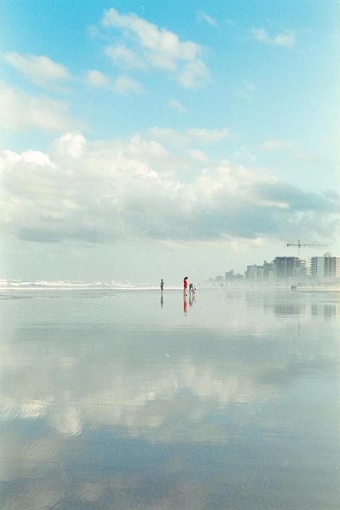 A group of people stand on a wet beach with buildings in the distance under a cloudy sky.