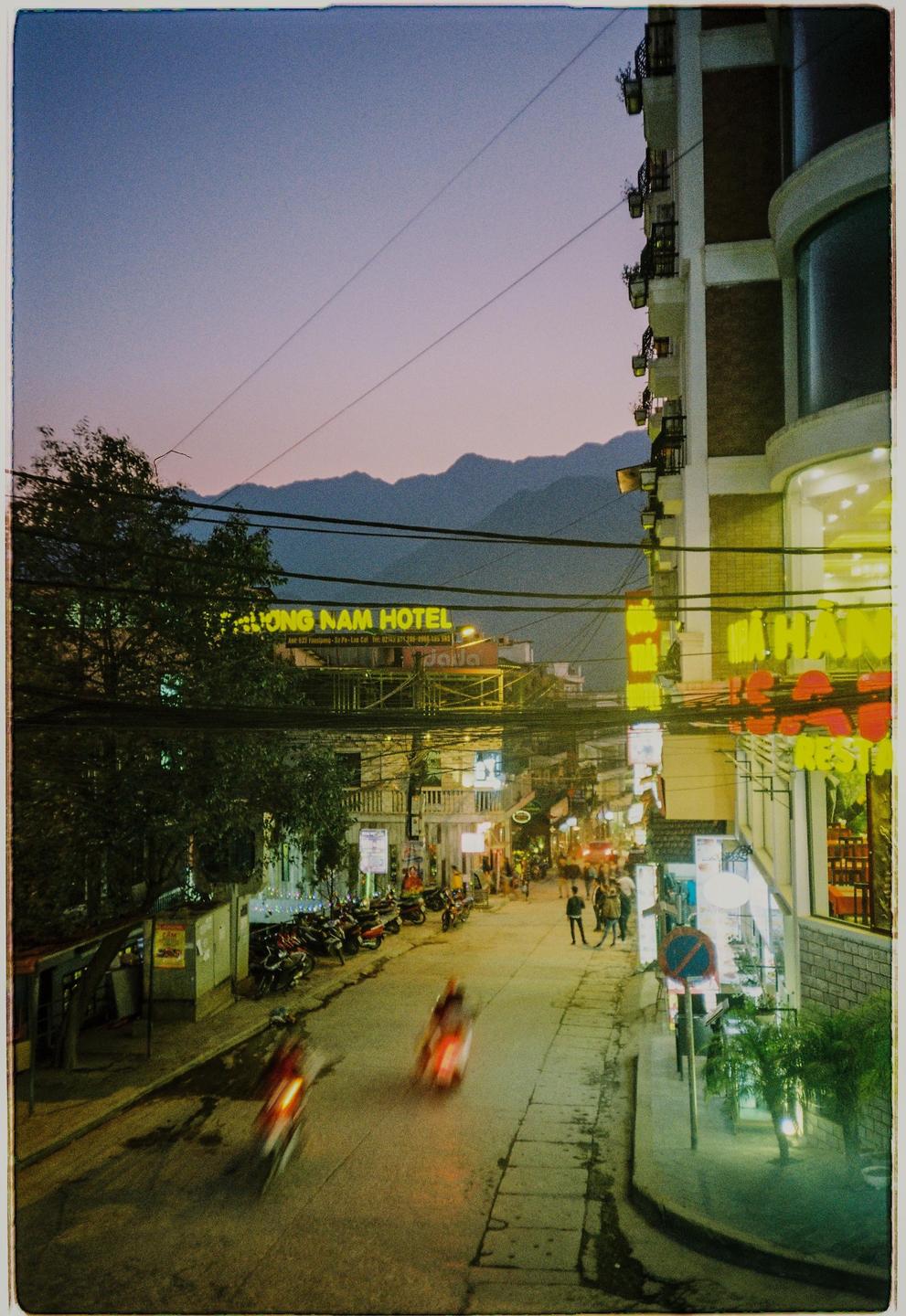 A street in SaPa Town is illuminated by neon signs and streetlights at dusk, with mountains in the background.