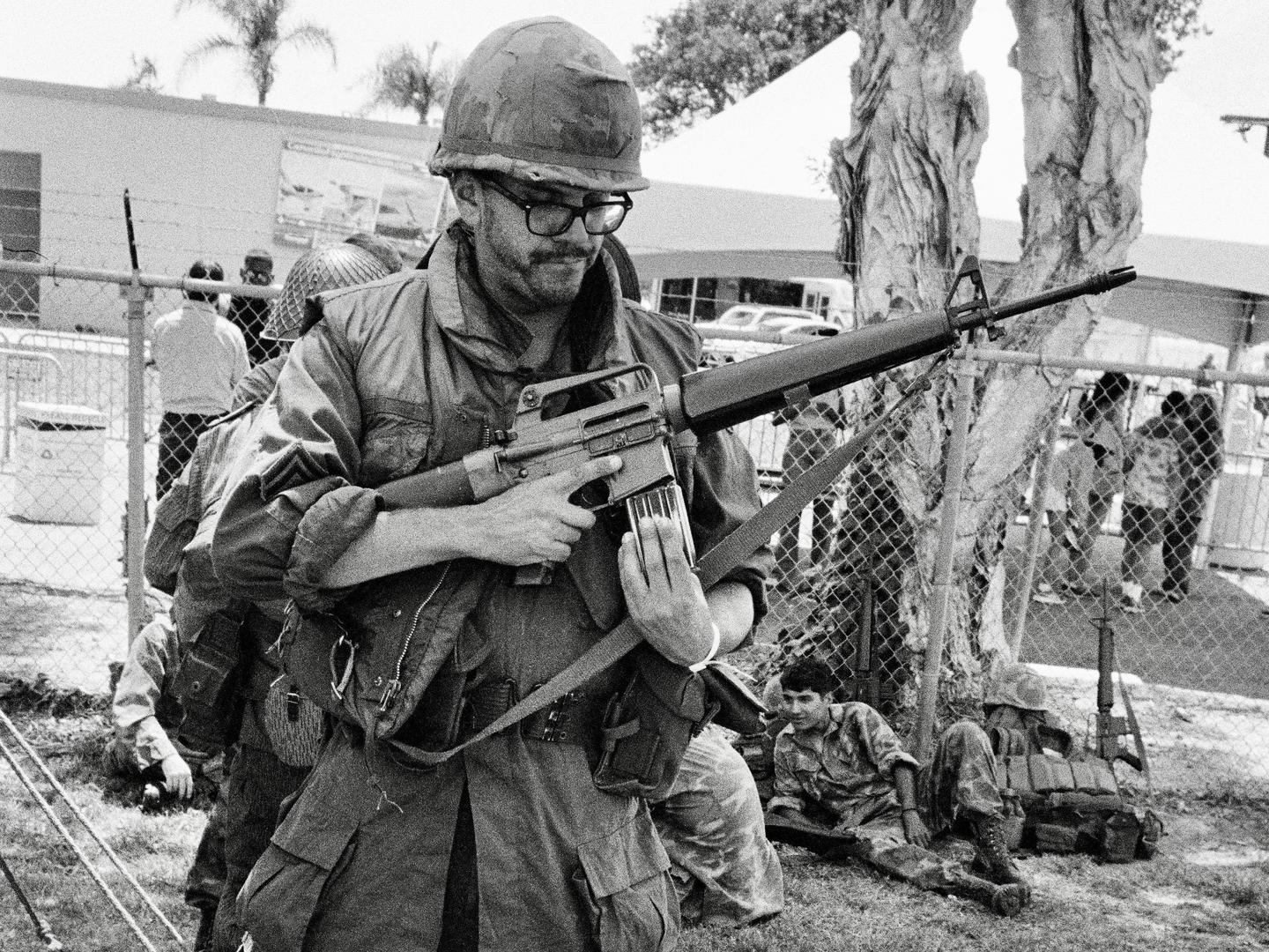 A man in military uniform and helmet holds a rifle while standing outdoors.