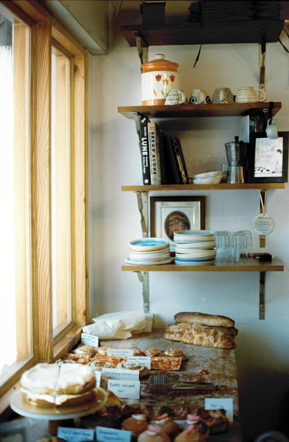 A variety of baked goods and pastries are displayed on a wooden counter next to a window and shelves with dishes and books.