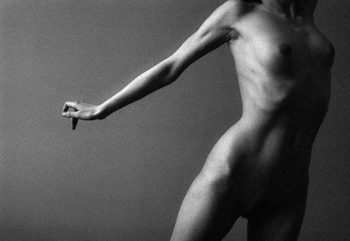 A nude woman is captured from the waist up, with her arm extended and fingers curled.
