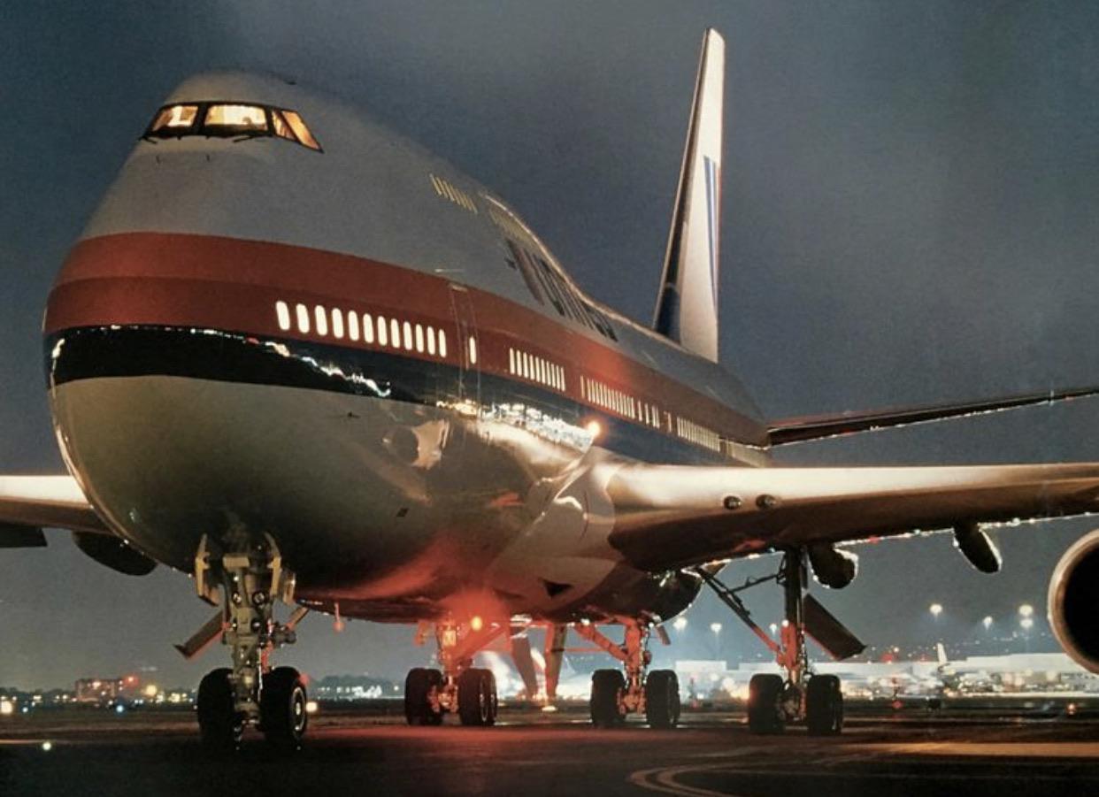A large airplane with red and blue stripes on its fuselage is parked on an airfield at night, illuminated by various lights.