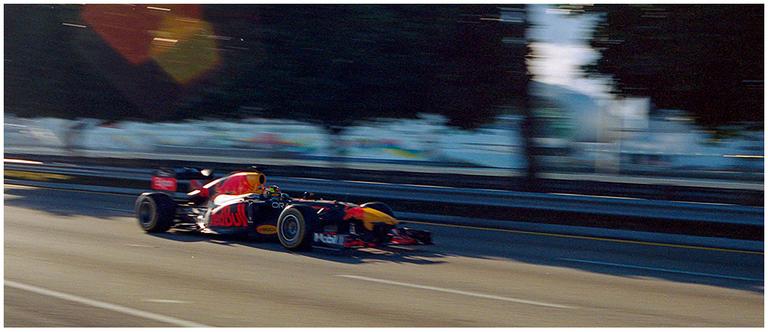 A red and black race car speeds down a track with blurred boats in the background.