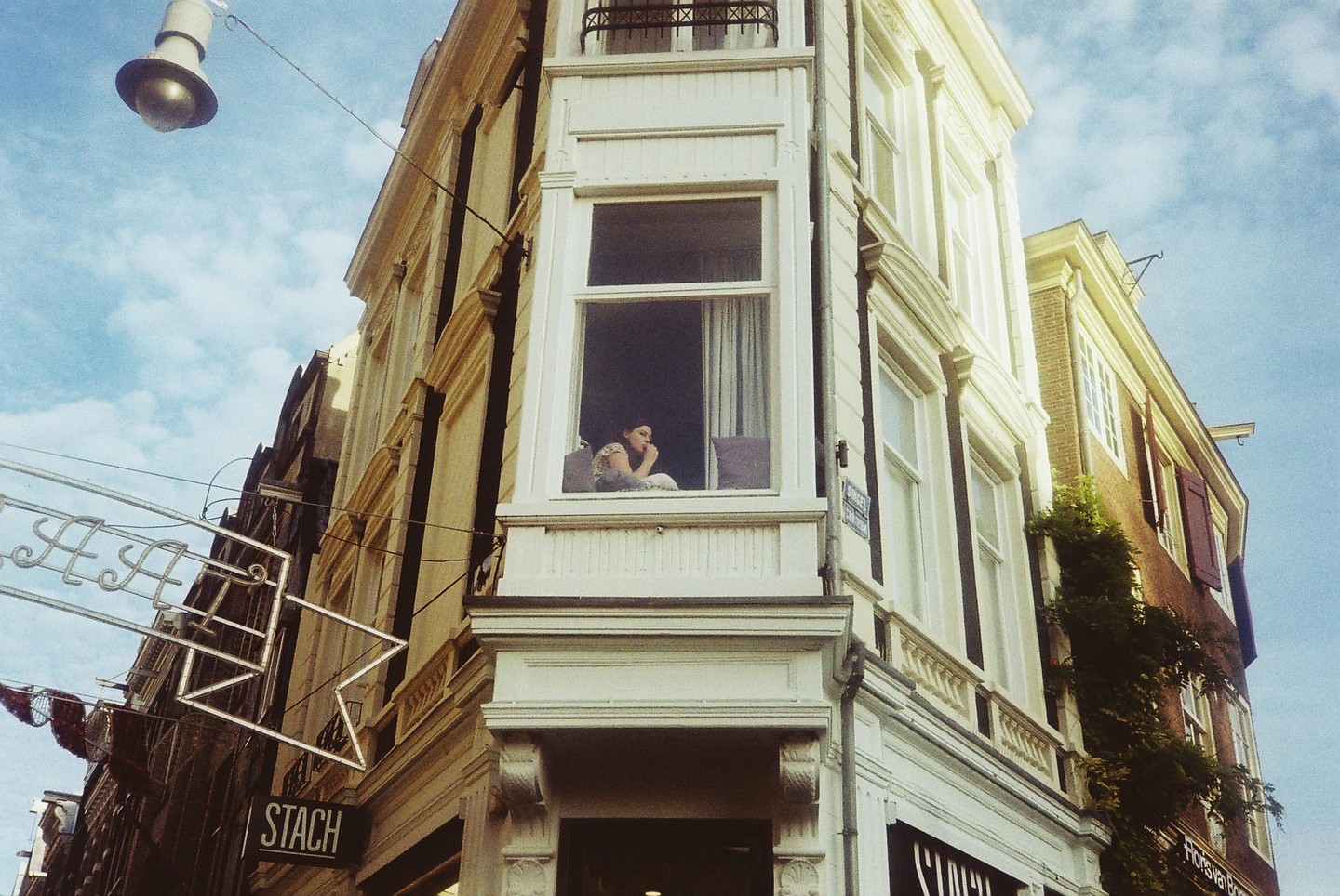 A woman sits in a bay window of a building with a street light and sign in the foreground.