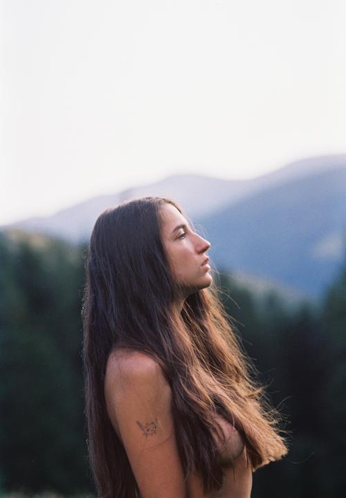 A woman with long hair and a tattoo on her arm looks up at the sky with mountains in the background.