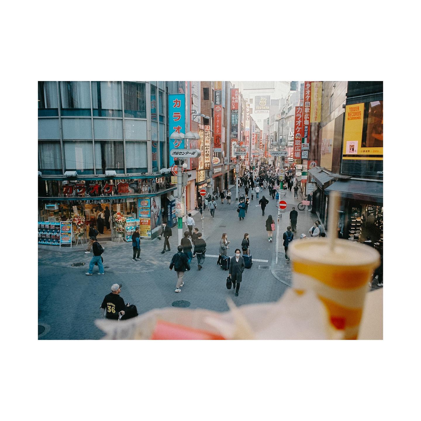 A busy street in Shibuya, Tokyo, is filled with pedestrians and neon signs, with a drink in the foreground.