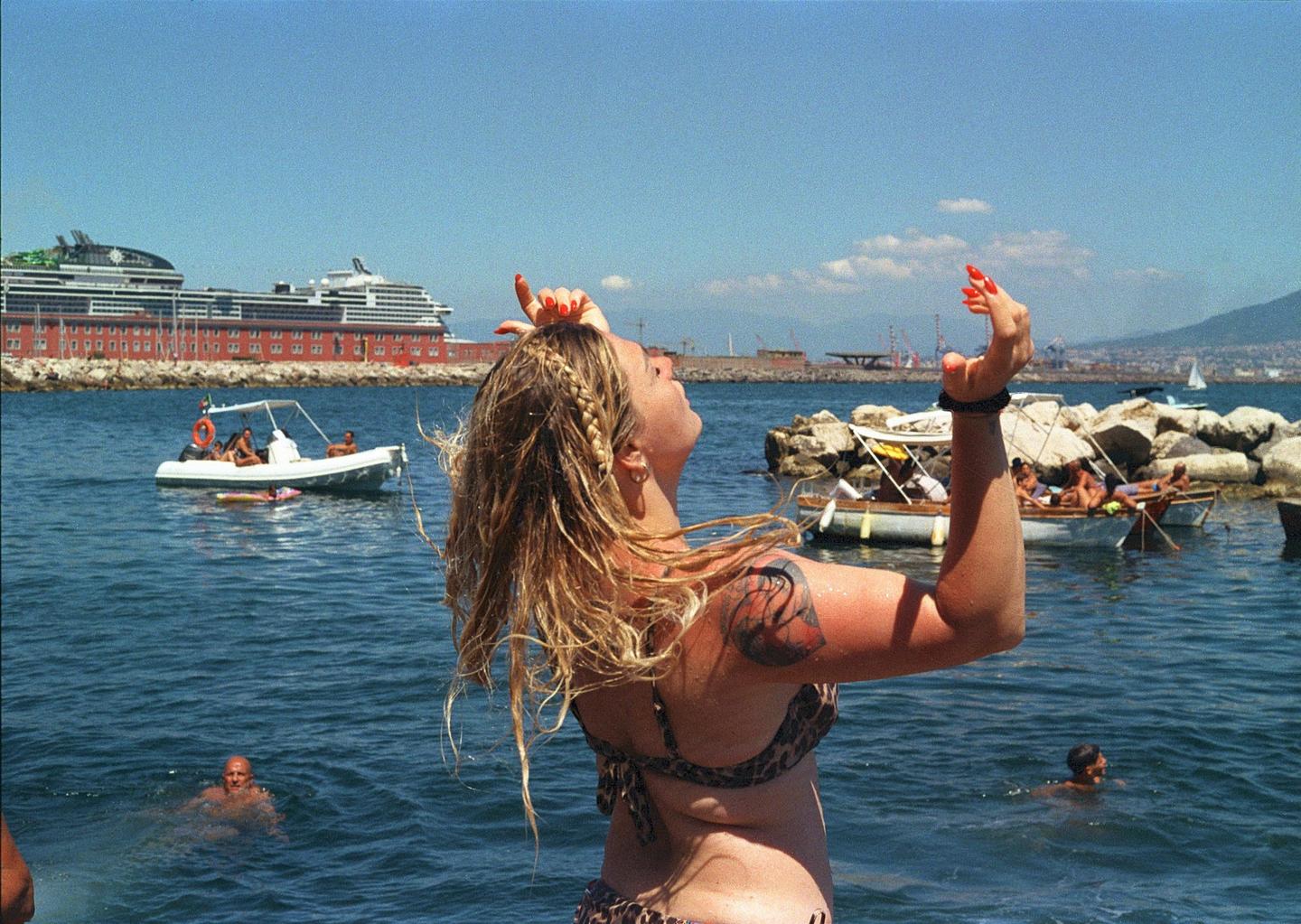 A woman with wet hair and a leopard print bikini stands in the water with her arms raised, looking up at the sky, with boats and a large ship in the background.