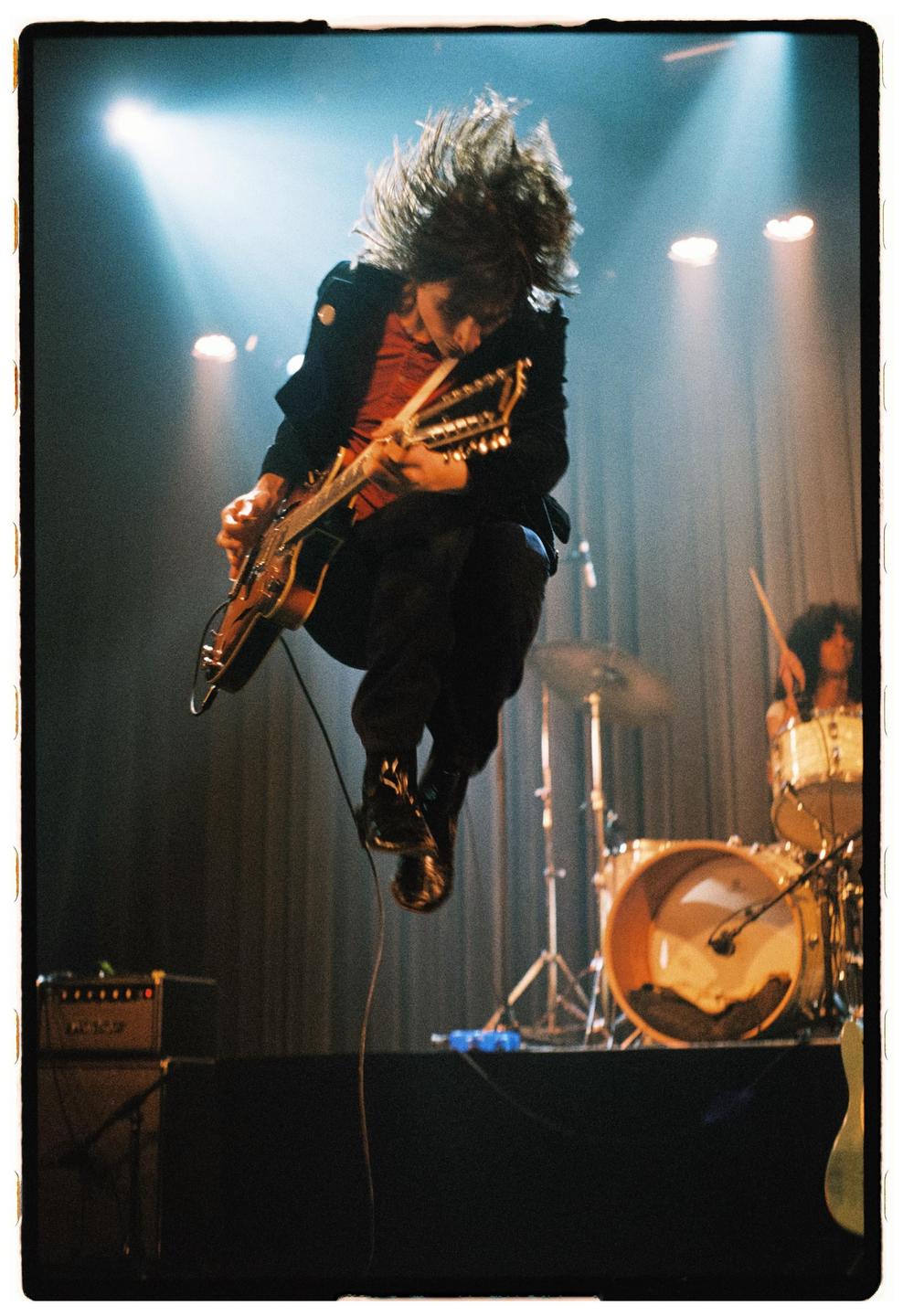 A musician with long hair jumps in the air while playing a guitar on stage, with a drummer visible in the background.