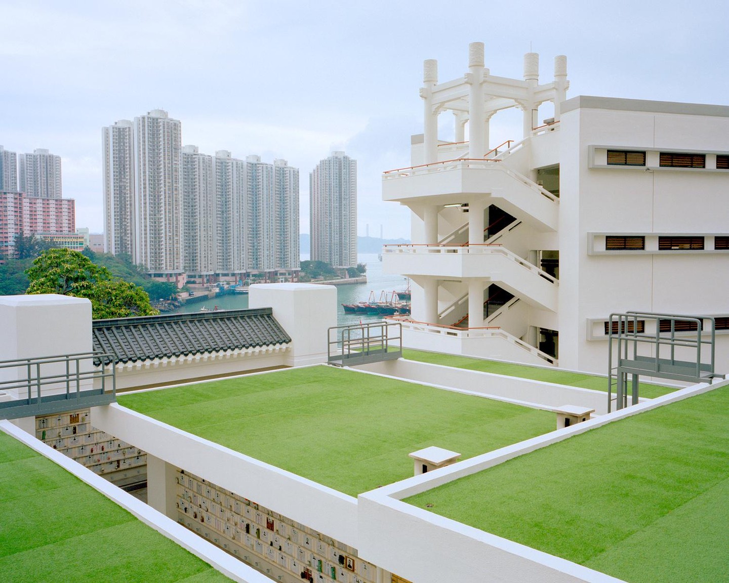 A modern white building with green turf roofs and a traditional dark tiled roof overlooks a bay with boats and tall buildings.
