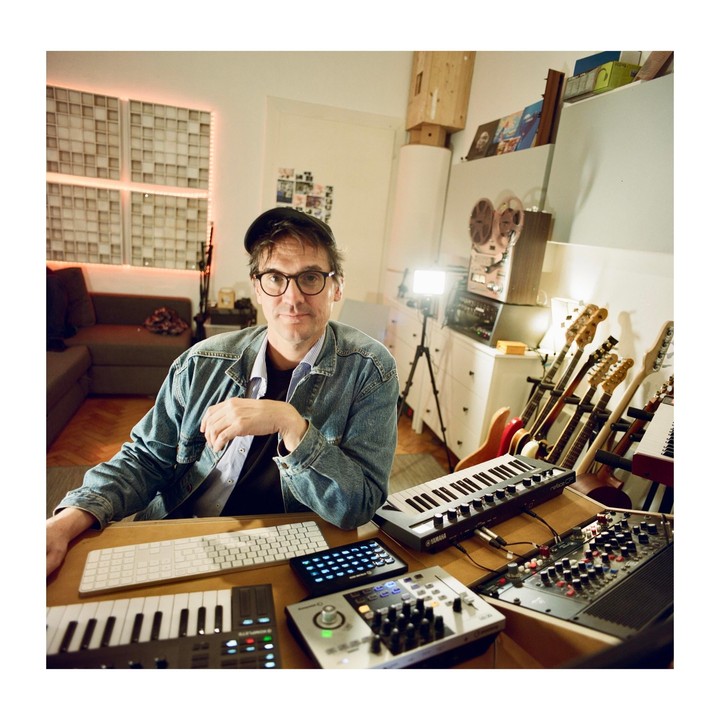A man wearing a denim jacket and a flat cap sits at a desk with musical instruments and recording equipment.
