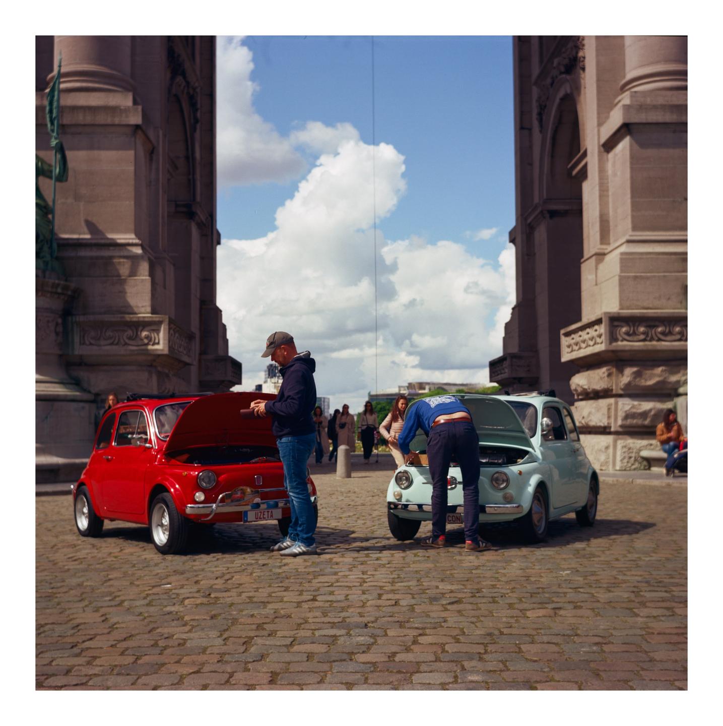Two men are working on small cars with their hoods open on a cobblestone street next to a large stone building.