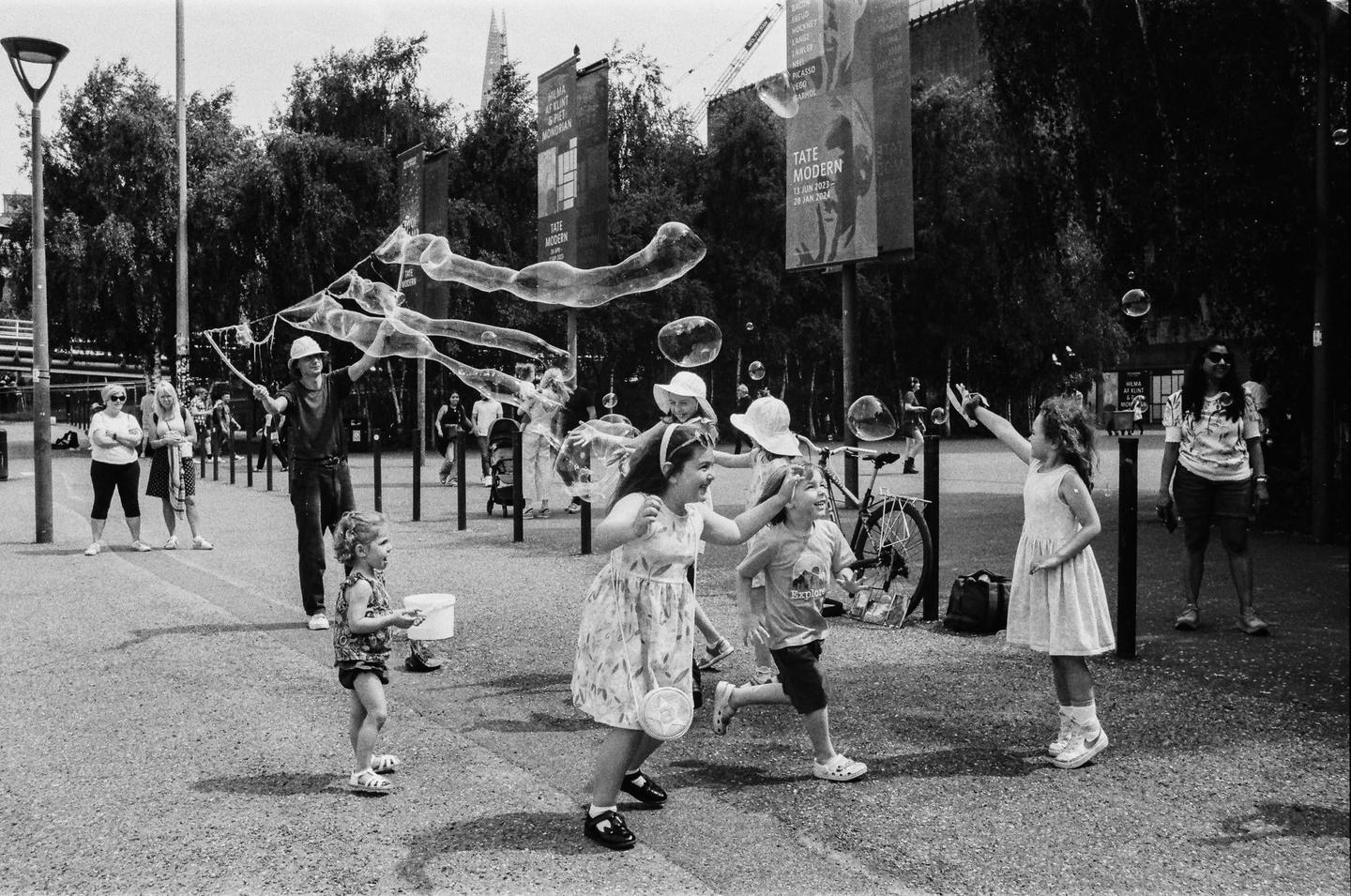 Children play with bubbles created by a man on a paved area with trees and buildings in the background.