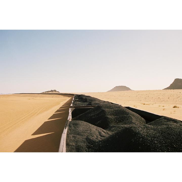 A train car filled with black rocks travels through a desert landscape with mountains in the distance.