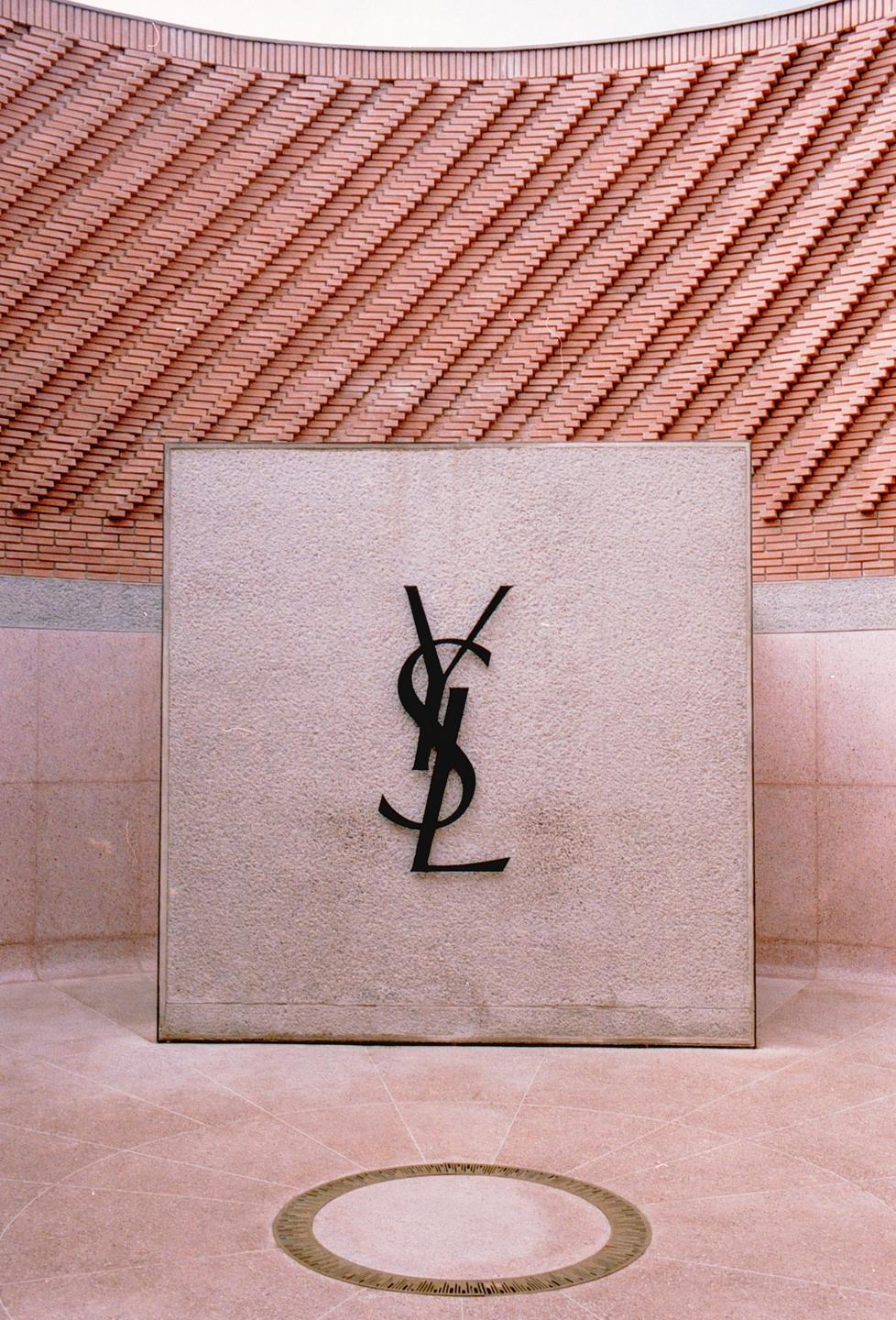 A large black Yves Saint Laurent logo is displayed on a light-colored stone slab in front of a curved brick wall.