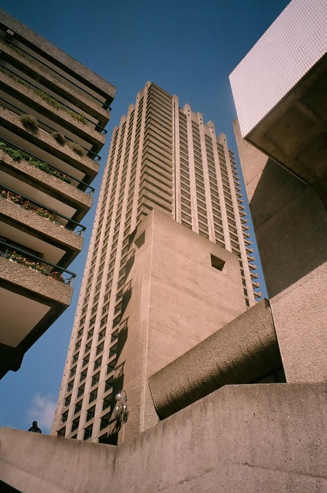 A low-angle view shows the brutalist architecture of the Barbican Estate in London, with a tall residential tower dominating the frame and balconies with plants visible on the left.