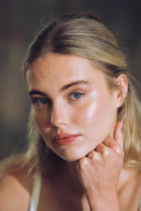 A woman with blonde hair and blue eyes looks directly at the viewer, resting her chin on her hand.