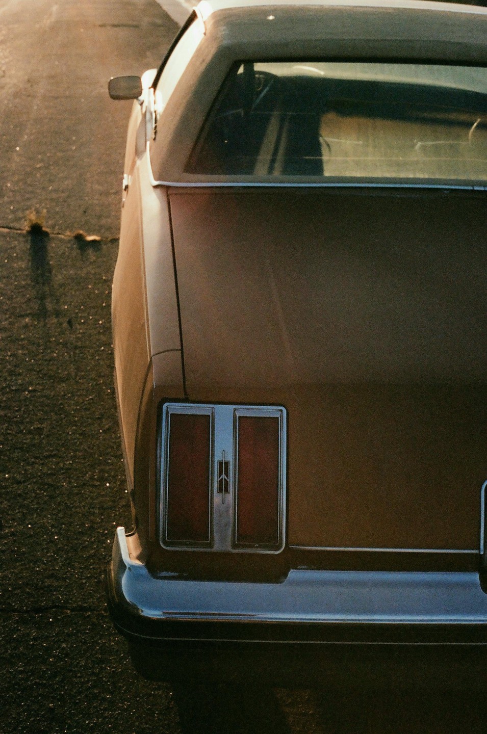 The rear of a brown car with prominent taillights and a chrome bumper is parked on an asphalt surface.