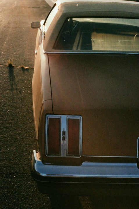 The rear of a brown car with prominent taillights and a chrome bumper is parked on an asphalt surface.