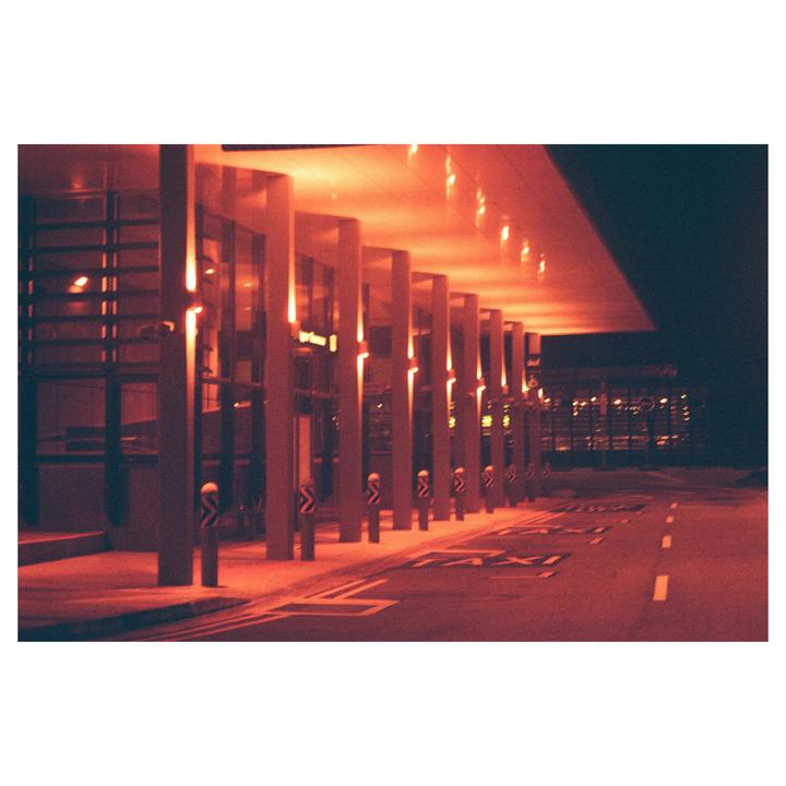 A long building with many pillars is illuminated by red light at night, with a road and "TAXI" painted on it in the foreground.