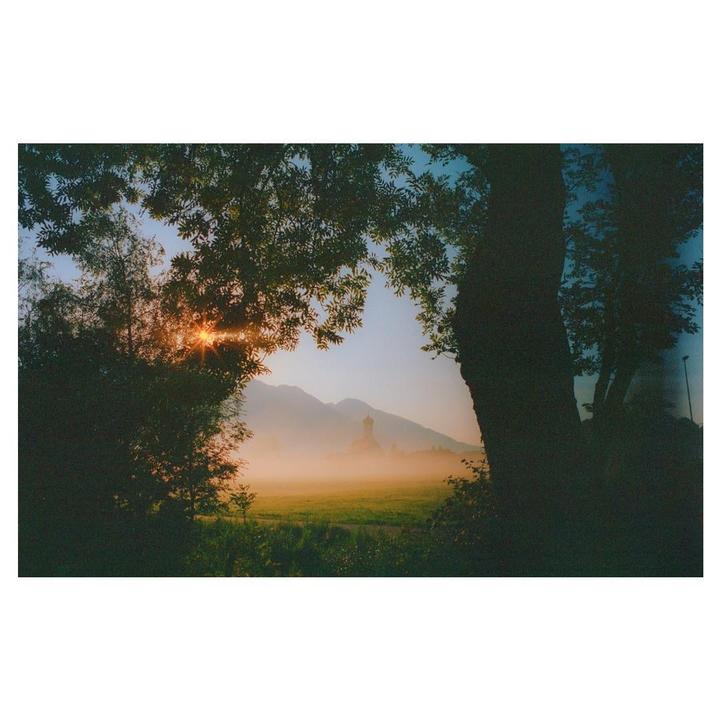 The sun shines through trees onto a hazy field with a church and mountains in the distance.