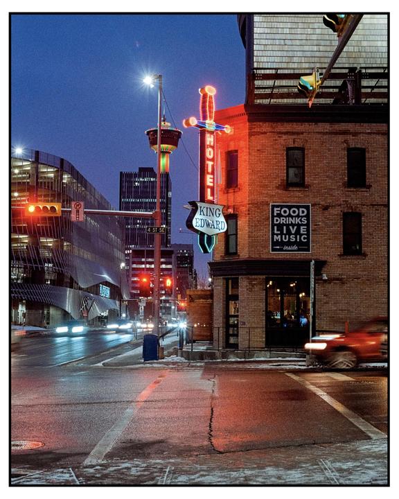 A neon sign for the King Edward Hotel glows brightly on a brick building at dusk, with car light trails on the street.