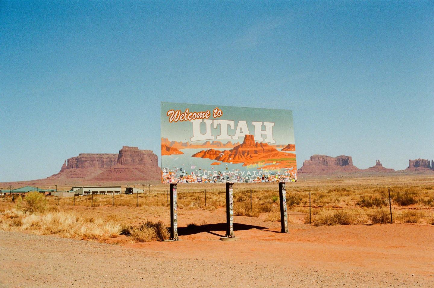 A "Welcome to Utah" sign stands in a desert landscape with mesas and buttes under a clear sky.