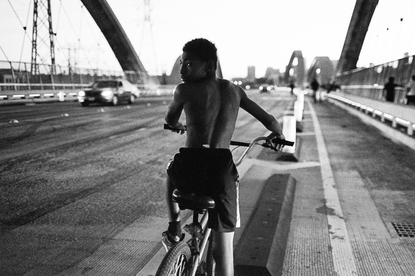 A boy on a bicycle looks over his shoulder on a bridge with cars and buildings in the background.