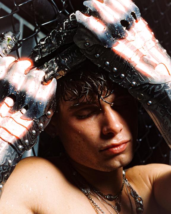 A man with wet hair and skin wears metal gauntlets and multiple necklaces, with his eyes closed.