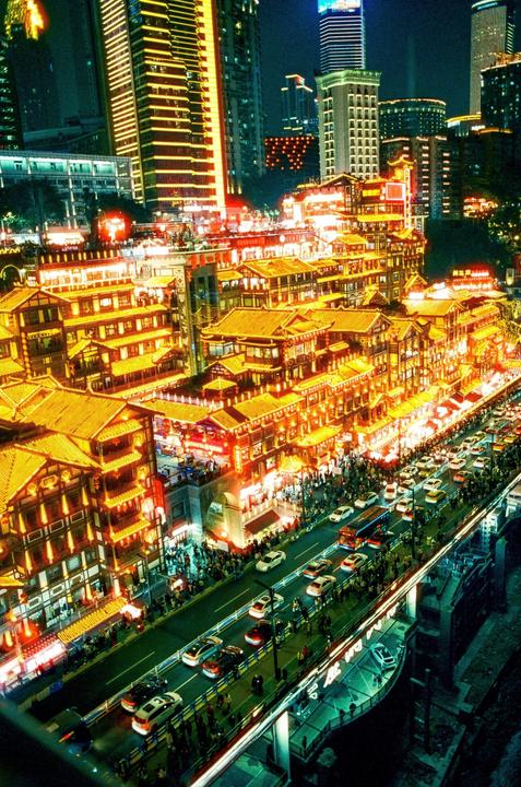 A busy street at night is lined with traditional buildings illuminated by bright orange and red lights, with modern skyscrapers in the background.