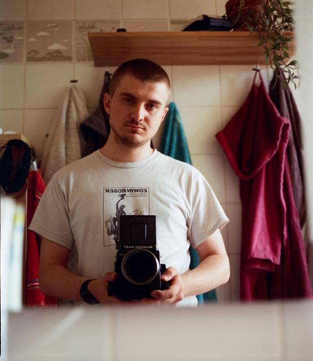 A man with a buzz cut and a white t-shirt holds a camera in a mirror self-portrait.
