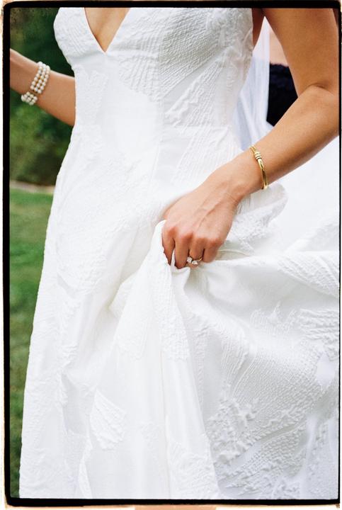 A woman in a white wedding dress holds up the skirt of her dress, revealing a ring on her finger and bracelets on her wrists.
