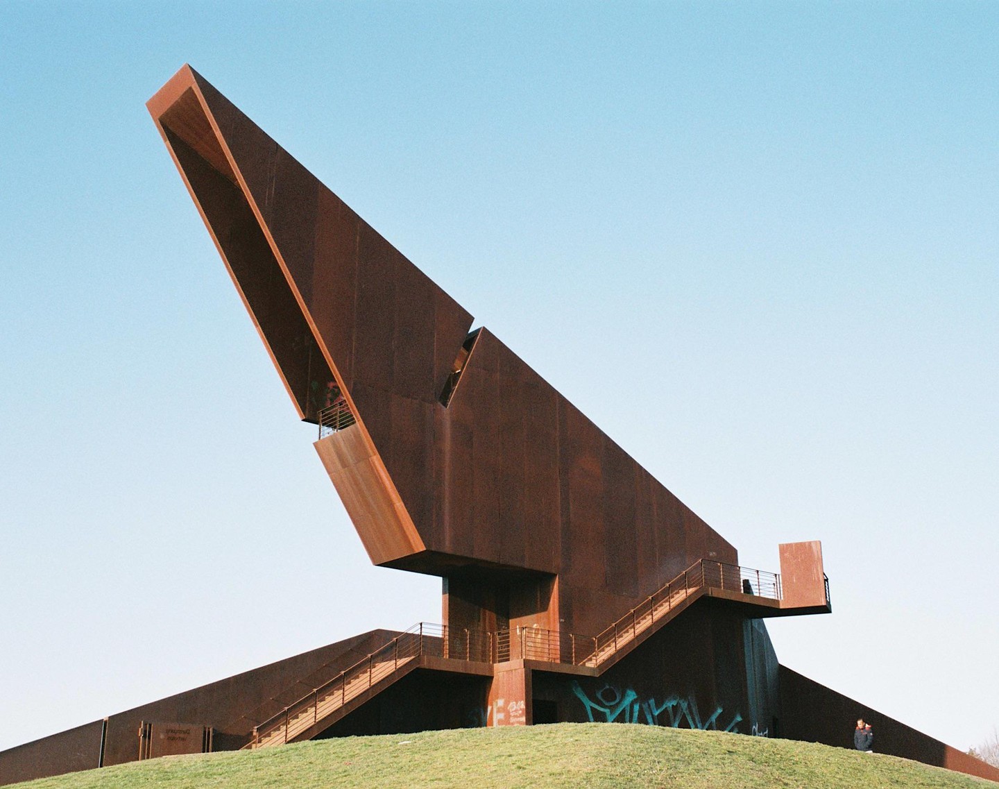 A large, rust-colored, angular building with stairs sits on a grassy hill under a clear sky.