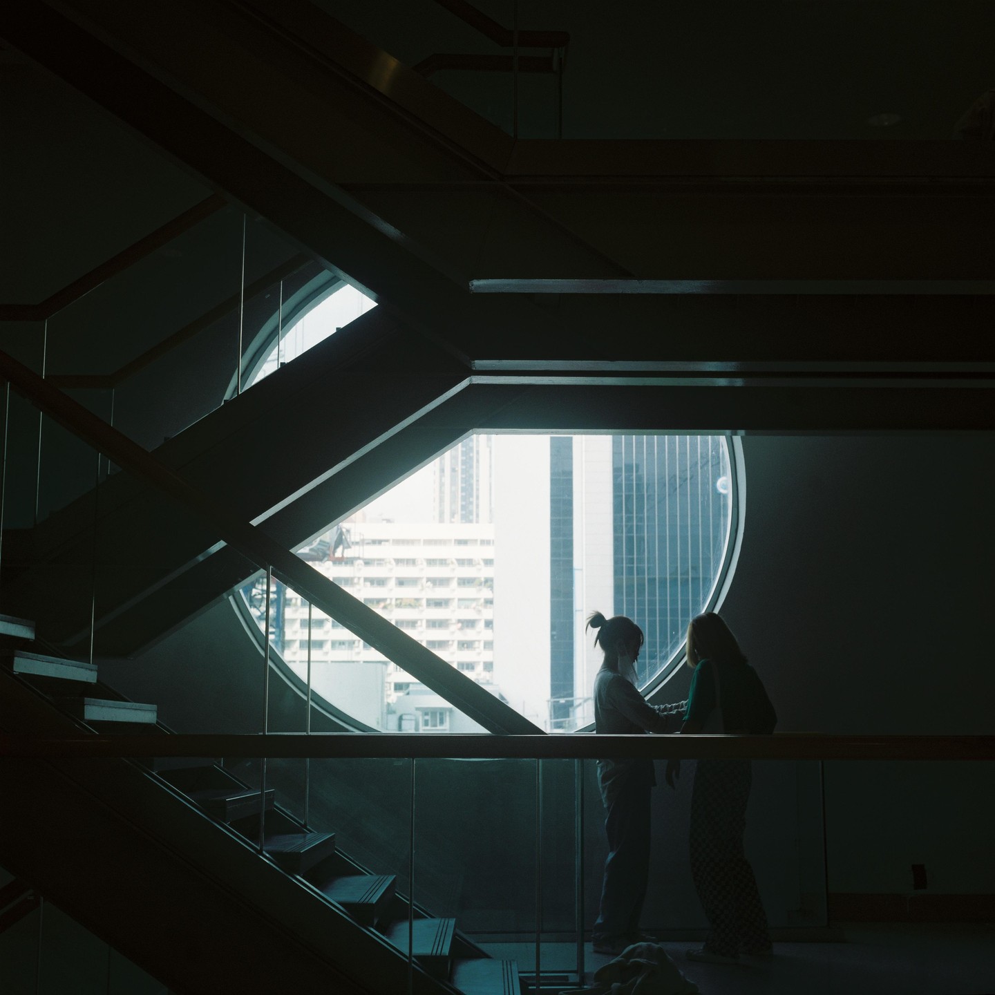 Two women stand silhouetted against a large circular window overlooking a city.