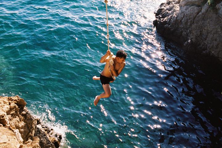 A man swings on a rope over sparkling blue water next to a rocky cliff.
