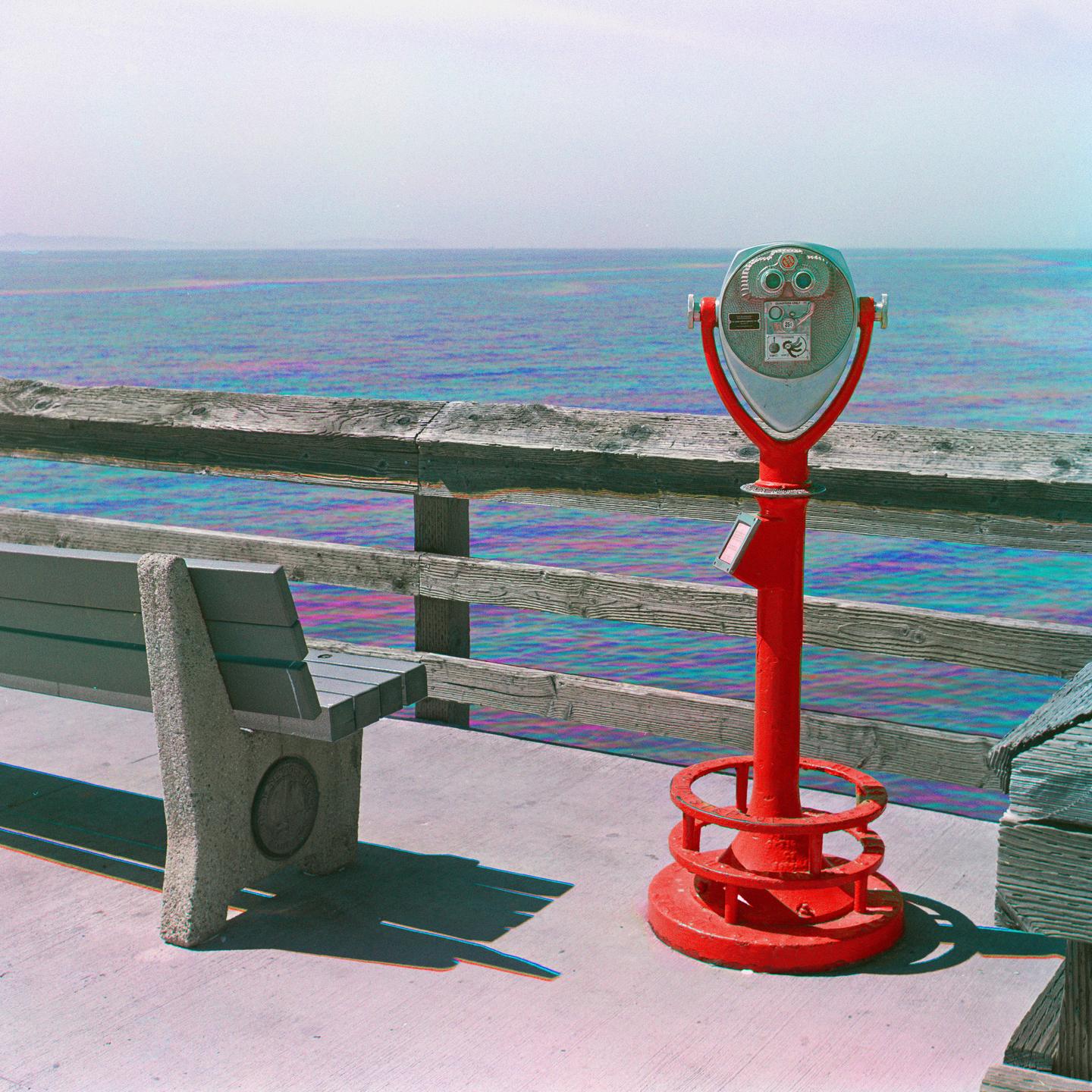 A red coin-operated binocular viewer stands on a pier overlooking the ocean next to a wooden bench.