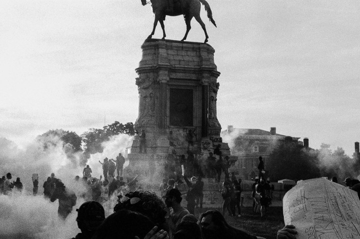 A crowd of people gathers around a large equestrian statue on a tall pedestal, with smoke filling the air.