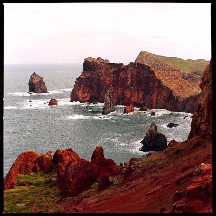 A dramatic coastline with red cliffs and rock formations juts into the ocean under a pale sky.