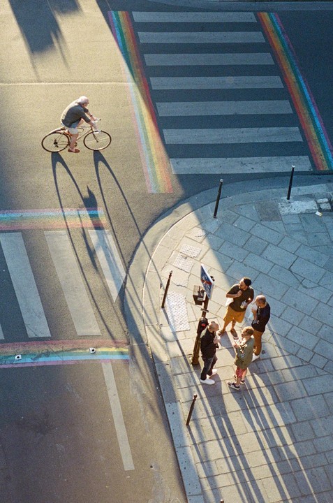 A high-angle view shows a man on a bicycle riding past a rainbow crosswalk while four people stand on the sidewalk.