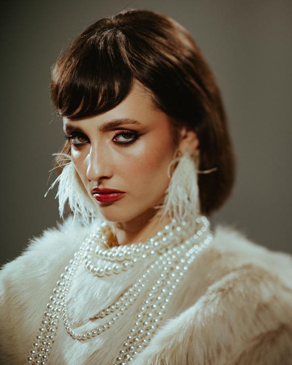 A woman with dark hair, red lipstick, and dramatic eye makeup wears a white fur coat, multiple strands of pearls, and feather earrings.