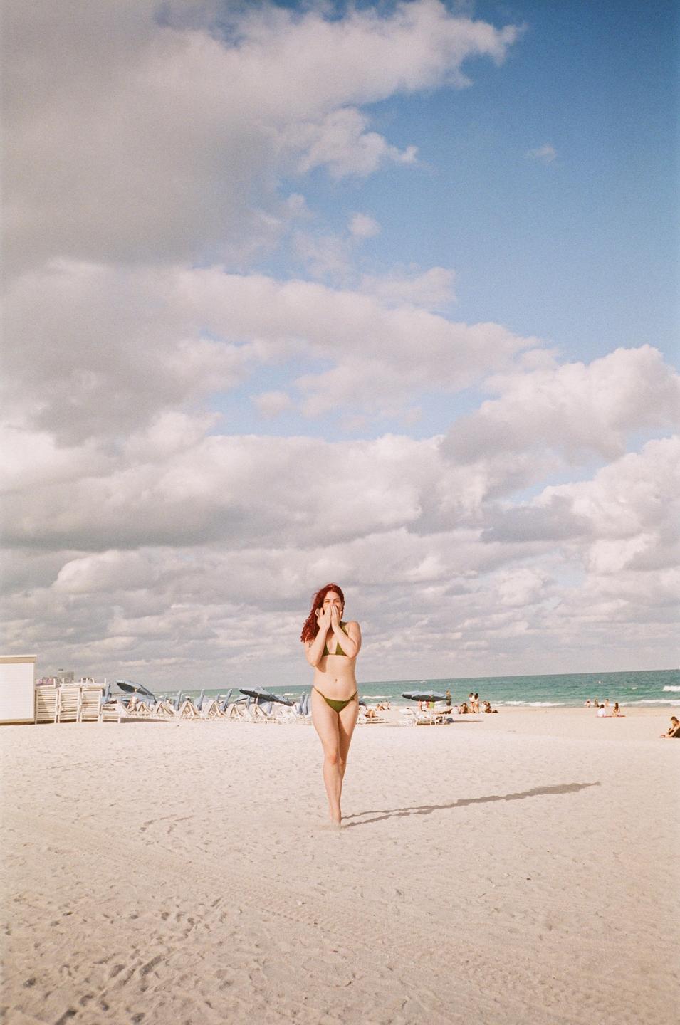 A woman in a bikini stands on a sandy beach with her hands covering her mouth.