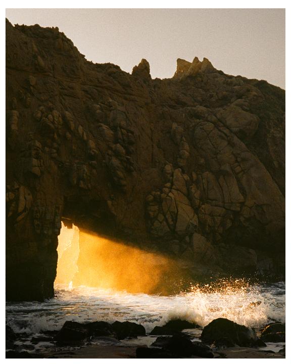 A beam of golden light shines through a rock archway onto crashing waves on a beach.