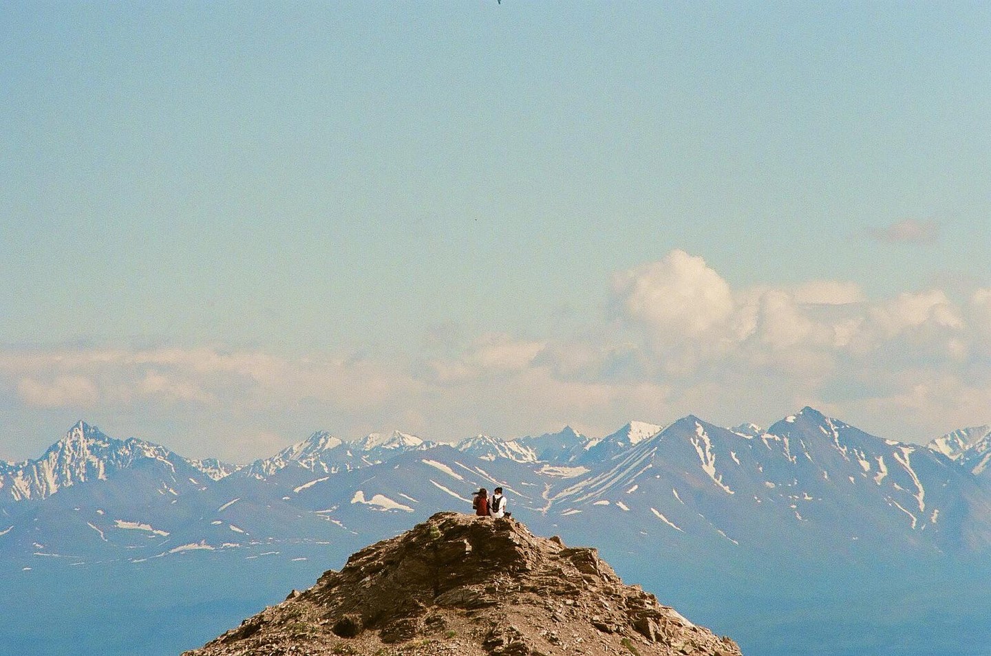 A couple sits on a rocky mountain peak overlooking a vast mountain range with snow-capped peaks under a clear sky.