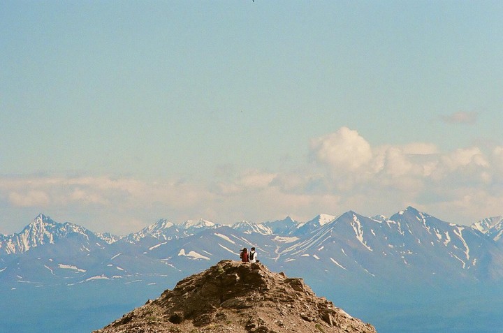 A couple sits on a rocky mountain peak overlooking a vast mountain range with snow-capped peaks under a clear sky.