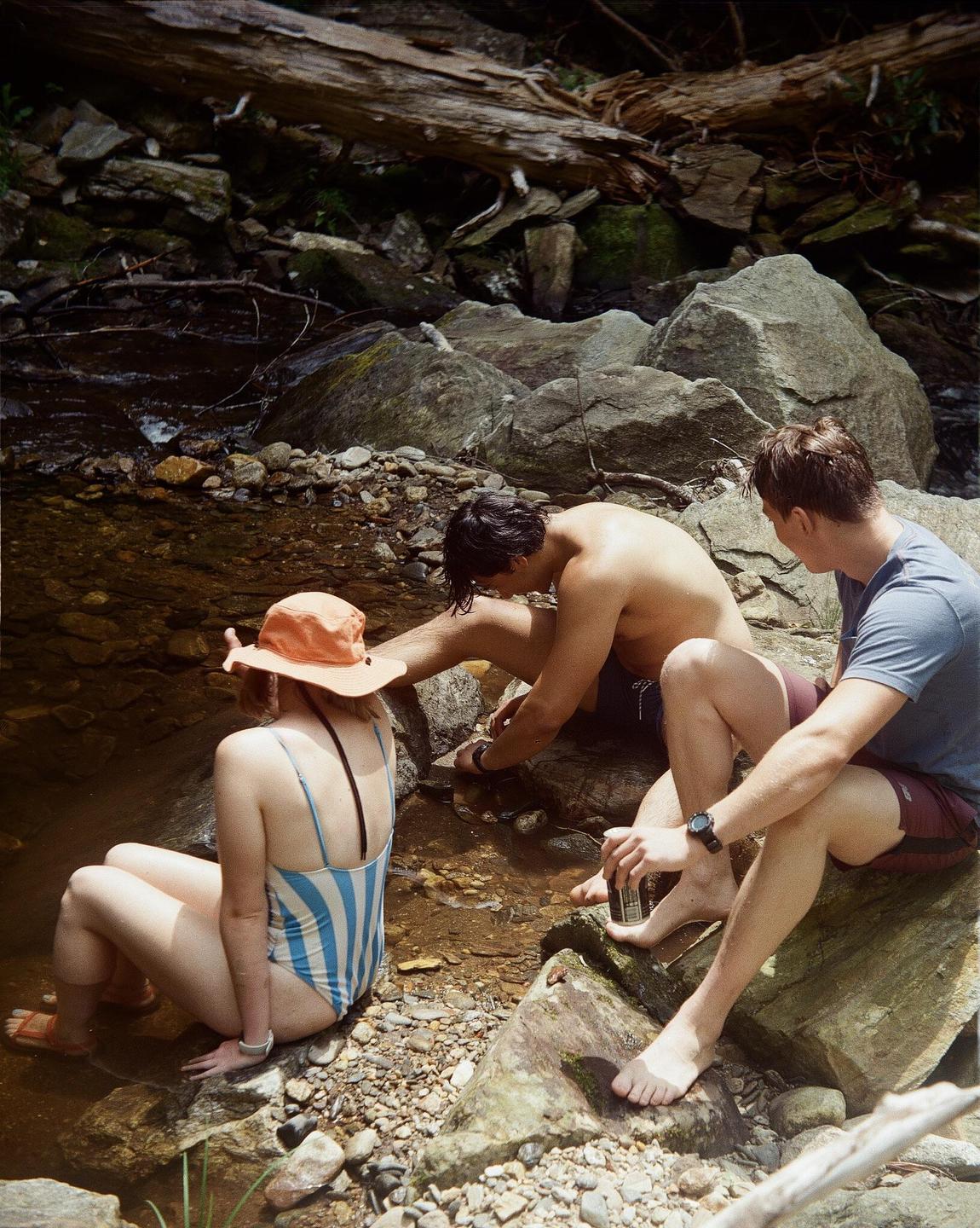 Three people sit on rocks by a stream with a fallen log in the background.