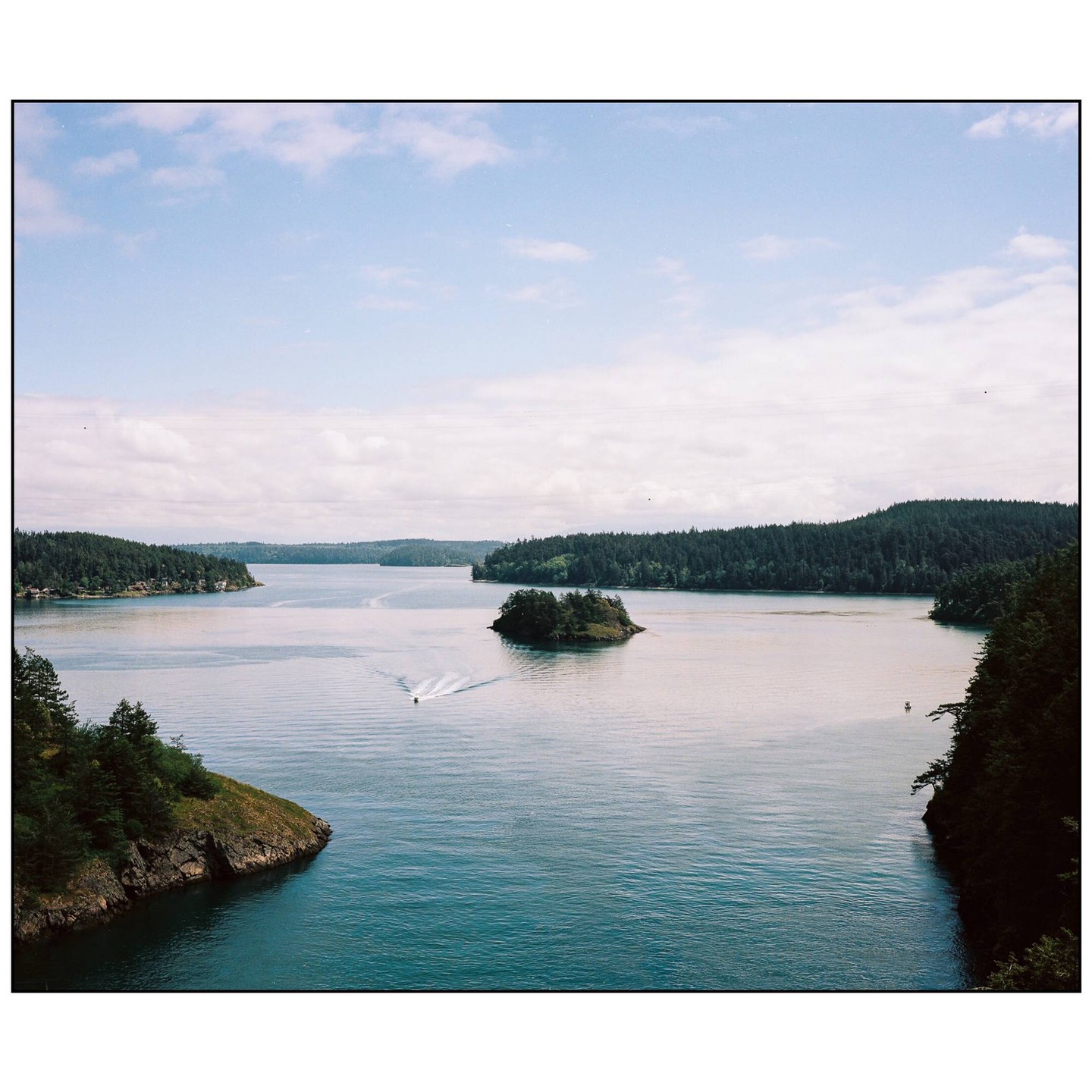 A small boat leaves a wake as it travels through a body of water surrounded by forested land and a small island.
