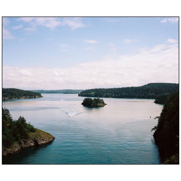 A small boat leaves a wake as it travels through a body of water surrounded by forested land and a small island.