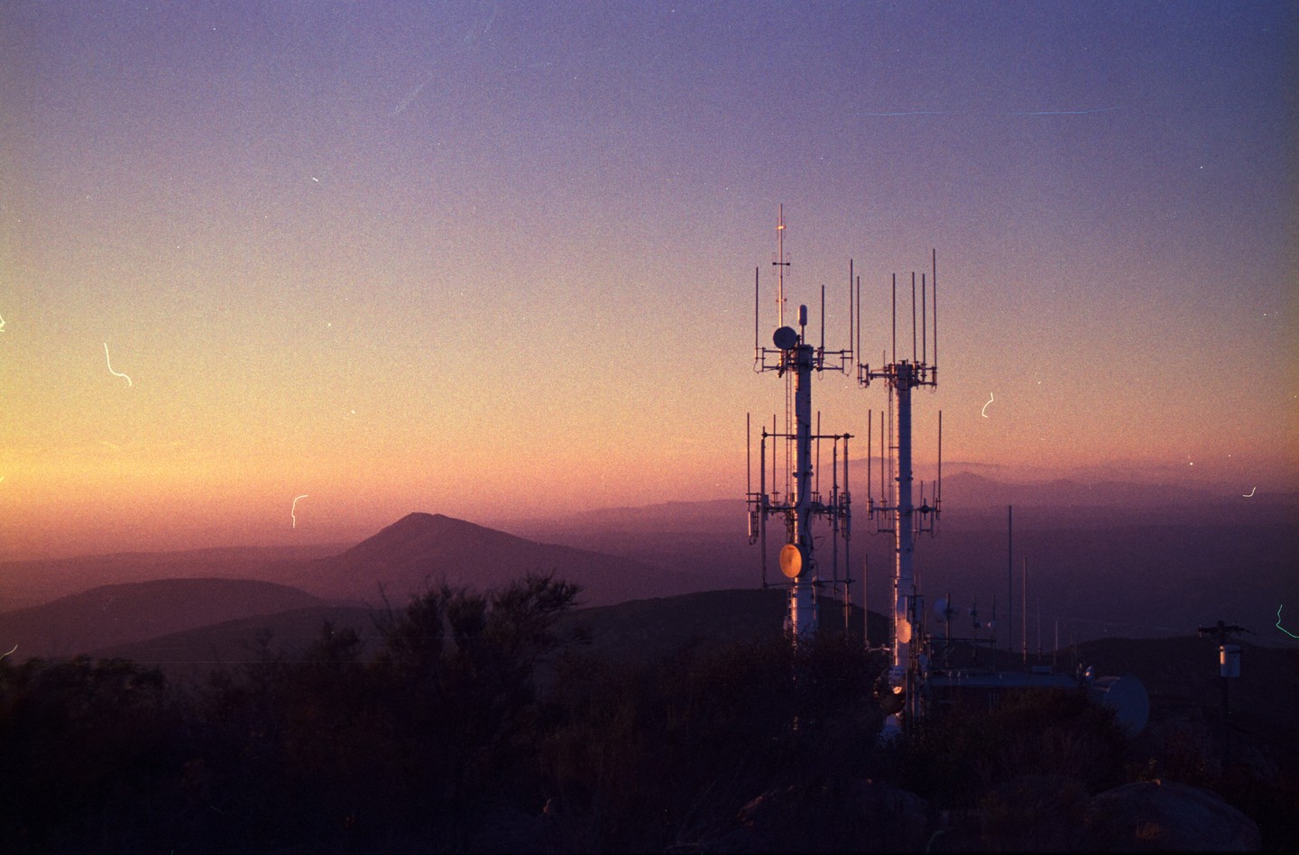 Two communication towers stand on a hill overlooking a mountain range at sunset.